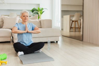 Yoga farkındalığı meditasyonu. Yetişkin bir kadın evde yoga yapıyor. Orta yaşlı kadın nilüfer çiçeğinde oturmuş yoga minderinde meditasyon yaparken poz veriyor. Yaşlı orta yaşlı kadın nefes egzersizi yapıyor.