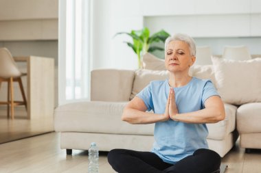 Yoga farkındalığı meditasyonu. Yetişkin bir kadın evde yoga yapıyor. Orta yaşlı kadın nilüfer çiçeğinde oturmuş yoga minderinde meditasyon yaparken poz veriyor. Yaşlı orta yaşlı kadın nefes egzersizi yapıyor.