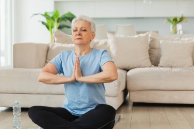 Yoga farkındalığı meditasyonu. Yetişkin bir kadın evde yoga yapıyor. Orta yaşlı kadın nilüfer çiçeğinde oturmuş yoga minderinde meditasyon yaparken poz veriyor. Yaşlı orta yaşlı kadın nefes egzersizi yapıyor.