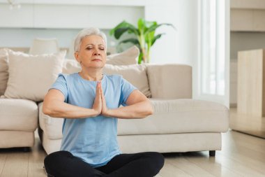 Yoga farkındalığı meditasyonu. Yetişkin bir kadın evde yoga yapıyor. Orta yaşlı kadın nilüfer çiçeğinde oturmuş yoga minderinde meditasyon yaparken poz veriyor. Yaşlı orta yaşlı kadın nefes egzersizi yapıyor.
