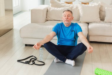 Yoga farkındalığı meditasyonu. Yetişkin bir adam evde yoga yapıyor. Orta yaşlı büyükbaba nilüfer çiçeğinde oturmuş yoga minderinde meditasyon yaparken poz veriyor. Yaşlı adam nefes alma pratiği yapıyor.