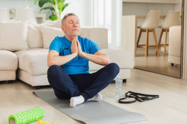 Yoga farkındalığı meditasyonu. Yetişkin bir adam evde yoga yapıyor. Orta yaşlı büyükbaba nilüfer çiçeğinde oturmuş yoga minderinde meditasyon yaparken poz veriyor. Yaşlı adam nefes alma pratiği yapıyor.