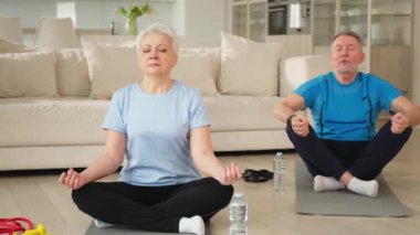 Yoga farkındalığı meditasyonu. Yetişkin bir çift evde yoga yapıyor. Orta yaşlı koca karısı nilüferde oturmuş yoga minderinde meditasyon yaparken poz veriyor. Aile nefes egzersizi yapıyor.