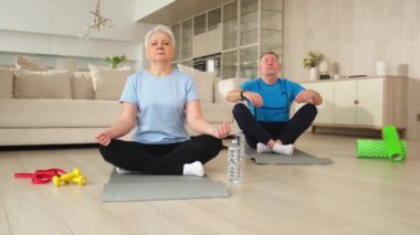 Yoga farkındalığı meditasyonu. Yetişkin bir çift evde yoga yapıyor. Orta yaşlı koca karısı nilüferde oturmuş yoga minderinde meditasyon yaparken poz veriyor. Aile nefes egzersizi yapıyor.