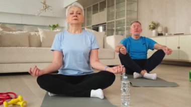 Yoga farkındalığı meditasyonu. Yetişkin bir çift evde yoga yapıyor. Orta yaşlı koca karısı nilüferde oturmuş yoga minderinde meditasyon yaparken poz veriyor. Aile nefes egzersizi yapıyor.