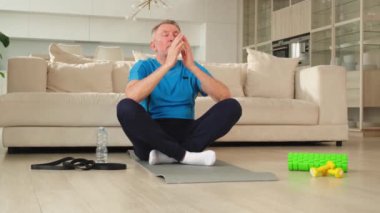 Yoga farkındalığı meditasyonu. Yetişkin bir adam evde yoga yapıyor. Orta yaşlı büyükbaba nilüfer çiçeğinde oturmuş yoga minderinde meditasyon yaparken poz veriyor. Yaşlı adam nefes alma pratiği yapıyor.
