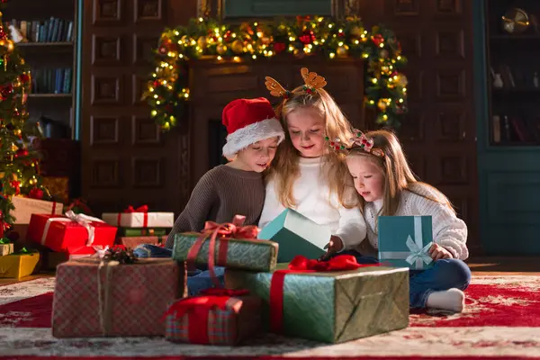 Mutlu Noeller Noel tatili. Çocuklar Noel ağacının yanındaki hediye kutusunu açıyor. Oturma odasında Noel ağacı şöminesi olan çocuklar sürpriz yüzlü hediye kutusunu açıyorlar. Evde Noel arifesi