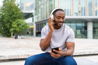 Müzik şarkısı konsepti. İyi bir başlangıç günü. Şehirde modern hit şarkıları dinleyen mutlu Afrikalı Amerikalı adam. Kulaklık takan adam telefonda en sevdiği müziği dinlemeyi sever. İnsanların yaşam tarzı neşesi