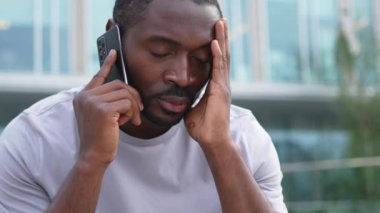 Üzgün, yalnız, hayal kırıklığına uğramış Afrikalı bir adam şehir caddesinde tek başına akıllı telefondan konuşuyor. Üzüntüden kötü haberler alan mutsuz bir insan cep telefonuyla telefona cevap veriyor. Stresli adam.