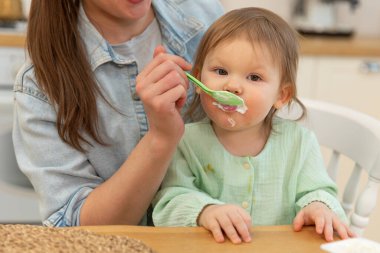 Evde mutlu bir aile. Anne mutfakta bebeğini kaşıkla besliyor. Dağınık suratlı küçük çocuk evde sağlıklı yiyecekler yiyor. Genç kadın anne, küçük kızına yemek veriyor.