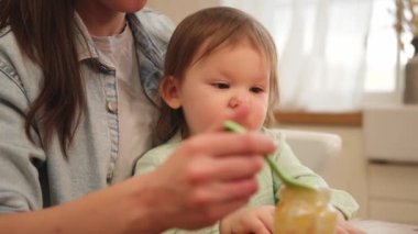 Evde mutlu bir aile. Anne mutfakta bebeğini kaşıkla besliyor. Dağınık suratlı küçük çocuk evde sağlıklı yiyecekler yiyor. Genç kadın anne, küçük kızına yemek veriyor.