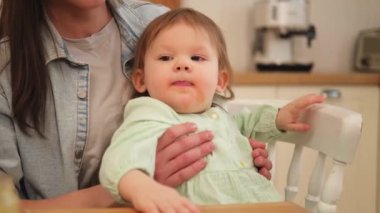 Evde mutlu bir aile. Anne mutfakta bebeğini kaşıkla besliyor. Dağınık suratlı küçük çocuk evde sağlıklı yiyecekler yiyor. Genç kadın anne, küçük kızına yemek veriyor.