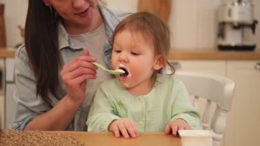 Evde mutlu bir aile. Anne mutfakta bebeğini kaşıkla besliyor. Dağınık suratlı küçük çocuk evde sağlıklı yiyecekler yiyor. Genç kadın anne, küçük kızına yemek veriyor.