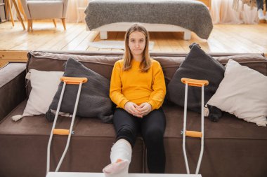 Kırık bacak. Kanepede oturan kırık bacaklı genç kız. Evdeki kırık bacağından iyileşen üzgün küçük çocuk. Alçı ve koltuk değneklerini rehabilitasyon için kullanan bir kız. Kaza yaralanması, kırık tedavisi.