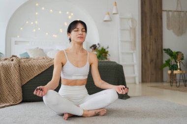 Yoga farkındalığı meditasyonu. Sağlıklı bir kadın evdeki oturma odasında yoga yapıyor. Lotus 'ta oturan kadın meditasyon yapıyor, gülümsüyor, evde dinleniyor. Nefes egzersizi yapan bir kız. Evde yoga.