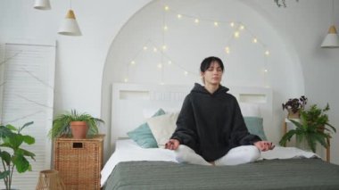 Yoga farkındalığı meditasyonu. Sağlıklı bir kadın yatak odasında yoga yapıyor. Nilüfer çiçekli bir kadın yatakta oturmuş, meditasyon yapıyor, gülümsüyor, evde dinleniyor. Nefes egzersizi yapan bir kız. Evde yoga.