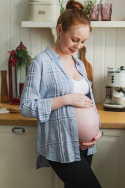 Hamilelik anneliği insanların gelecek beklentisi. Koca göbekli hamile bir kadın mutfakta gülümsüyor. Karnına sarılan ve hamileliğin tadını çıkaran bir kız. Annelik şefkati ebeveynlik yeni yaşam konsepti