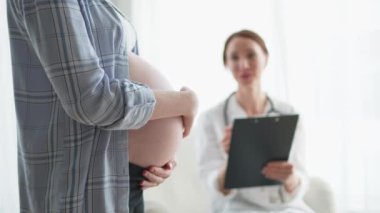Hamile kadın elleri doktor muayenehanesinde koca göbeğe dokunuyor. Kadın doktor hamile kadını muayene ediyor. Jinekolog doktor hastaya hastanede hamilelik konusunda danışmanlık yapıyor. Doktor kontrolüne git.