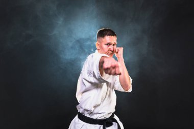 Siyah kuşaklı karate dövüş sanatları için beyaz kimonolu güçlü adam siyah arka plandaki dövüş eğitimine hazırlanıyor. Antrenman hareketleri yumruklar gölge dövüşü alıştırmaları antrenmana hazır.