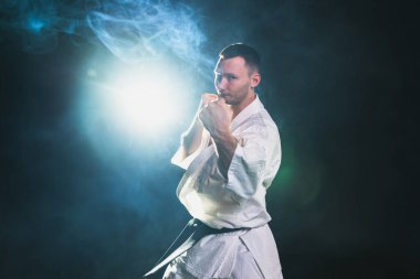 Siyah kuşaklı karate dövüş sanatları için beyaz kimonolu güçlü adam siyah arka plandaki dövüş eğitimine hazırlanıyor. Antrenman hareketleri yumruklar gölge dövüşü alıştırmaları antrenmana hazır.