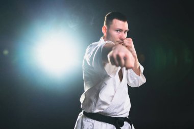 Siyah kuşaklı karate dövüş sanatları için beyaz kimonolu güçlü adam siyah arka plandaki dövüş eğitimine hazırlanıyor. Antrenman hareketleri yumruklar gölge dövüşü alıştırmaları antrenmana hazır.