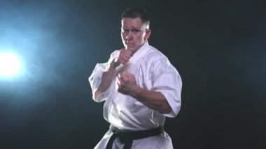 Siyah kuşaklı karate dövüş sanatları için beyaz kimonolu güçlü adam siyah arka plandaki dövüş eğitimine hazırlanıyor. Antrenman hareketleri yumruklar gölge dövüşü alıştırmaları antrenmana hazır.