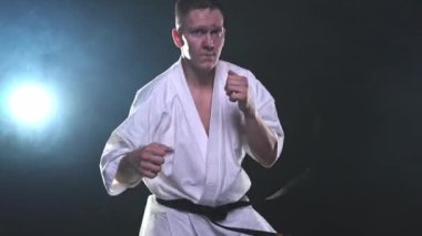 Siyah kuşaklı karate dövüş sanatları için beyaz kimonolu güçlü adam siyah arka plandaki dövüş eğitimine hazırlanıyor. Antrenman hareketleri yumruklar gölge dövüşü alıştırmaları antrenmana hazır.