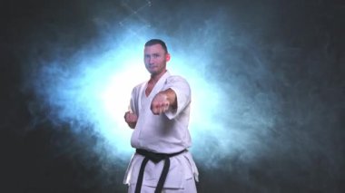 Siyah kuşaklı karate dövüş sanatları için beyaz kimonolu güçlü adam siyah arka plandaki dövüş eğitimine hazırlanıyor. Antrenman hareketleri yumruklar gölge dövüşü alıştırmaları antrenmana hazır.