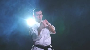 Siyah kuşaklı karate dövüş sanatları için beyaz kimonolu güçlü adam siyah arka plandaki dövüş eğitimine hazırlanıyor. Antrenman hareketleri yumruklar gölge dövüşü alıştırmaları antrenmana hazır.