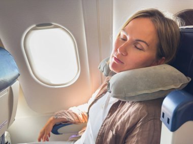Uçakta dinlen. Genç bir kadın seyahat için boyun yastığını kullanarak lomboz kenarında oturuyor. Uçakla uçan yolcu kız. Uçakta uyuyan kız tatile çıkıyor. Gezgin kız.