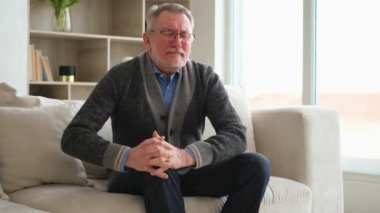Diz ağrısı, eklem iltihabı, vücut hastalıkları, sağlık hizmetleri. Evdeki kanepede oturan mutsuz, orta yaşlı, diz ağrısı çeken bir adam. Yaşlı büyükbaba bacağına dokunuyor ve dizinde ağrı hissediyor.