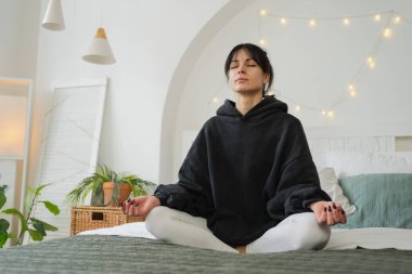 Yoga farkındalığı meditasyonu. Sağlıklı bir kadın yatak odasında yoga yapıyor. Nilüfer çiçekli bir kadın yatakta oturmuş, meditasyon yapıyor, gülümsüyor, evde dinleniyor. Nefes egzersizi yapan bir kız. Evde yoga.