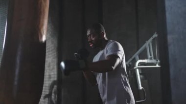 Beden eğitiminde spor. Güçlü Afrikalı dövüşçü boks torbasıyla yumruk eğitimi veriyor. Spor salonunda boks torbası yumruklayan sağlıklı bir boksör. Boks salonunda antrenman günü. Vücut çalıştırma eğitimine uygun güç