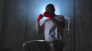 Beden eğitiminde spor. Afrikalı dövüşçü dövüşmeye hazırlanıyor kırmızı boksla yumruk atarken spor sargılarını spor salonunda sarar. Güçlü adam boks antrenmanına hazır yumrukları eğitiyor.