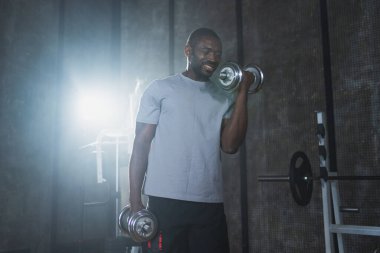 Beden eğitiminde spor. Spor salonunda dambılları toplayan Afrikalı Amerikalı bir vücut geliştirici. Fitness kulübünde halterli kasları çalıştırmak için ağırlık çalışıyorum. Spor yapan bir adam. Sağlıklı yaşam tarzı