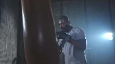 Beden eğitiminde spor. Güçlü Afrikalı dövüşçü boks torbasıyla yumruk eğitimi veriyor. Spor salonunda boks torbası yumruklayan sağlıklı bir boksör. Boks salonunda antrenman günü. Vücut çalıştırma eğitimine uygun güç