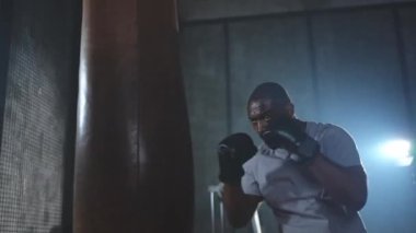 Beden eğitiminde spor. Güçlü Afrikalı dövüşçü boks torbasıyla yumruk eğitimi veriyor. Spor salonunda boks torbası yumruklayan sağlıklı bir boksör. Boks salonunda antrenman günü. Vücut çalıştırma eğitimine uygun güç