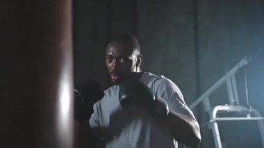 Beden eğitiminde spor. Güçlü Afrikalı dövüşçü boks torbasıyla yumruk eğitimi veriyor. Spor salonunda boks torbası yumruklayan sağlıklı bir boksör. Boks salonunda antrenman günü. Vücut çalıştırma eğitimine uygun güç