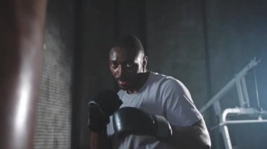Beden eğitiminde spor. Güçlü Afrikalı dövüşçü boks torbasıyla yumruk eğitimi veriyor. Spor salonunda boks torbası yumruklayan sağlıklı bir boksör. Boks salonunda antrenman günü. Vücut çalıştırma eğitimine uygun güç