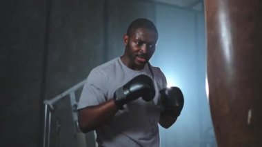 Beden eğitiminde spor. Güçlü Afrikalı dövüşçü boks torbasıyla yumruk eğitimi veriyor. Spor salonunda boks torbası yumruklayan sağlıklı bir boksör. Boks salonunda antrenman günü. Vücut çalıştırma eğitimine uygun güç