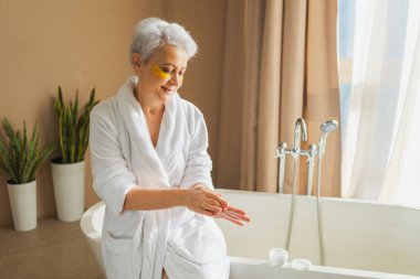 Olgun, yaşlı bir kadın ellerinde nemlendirici vücut losyonu sürüyor. Son sınıf öğrencisi kadın banyoda günlük cilt bakımını yaparken anti-yaş kremi sürüyor. Güzellik tedavisi vücut bakımını kapsar.