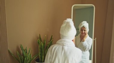 Yetişkin, yaşlı bir kadın aynaya bakıyor ve günlük güzellik kremi sürüyor. Cildinde beyaz maske kremi olan yaşlı bir kadın cilt bakımı görüyor.