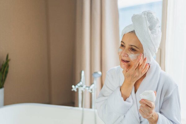 Mature adult old woman applying nourishing anti age face cream doing daily beauty routine. Senior woman with white mask or creme on skin gets facial treatment Skin care aging beauty skincare cosmetics