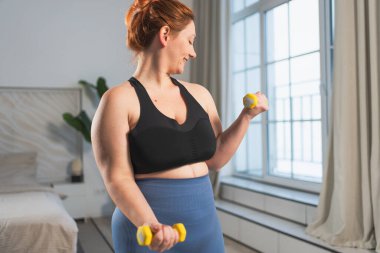 Fitness antrenmanı. Dambılları kaldıran kadın. Artı ev içinde spor egzersizi yapan beden bir kadın. Spor giyimli kız kilo vermek için ağırlık çalışması yapıyor. Spor ve zindelik