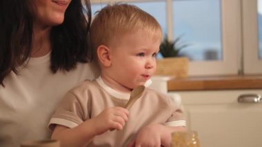 Evde mutlu bir aile. Anne, bebeğini mutfakta kaşıkla besliyor. Dağınık suratlı küçük çocuk evde sağlıklı yiyecekler yiyor. Genç kadın anne oğluna yemek veriyor.
