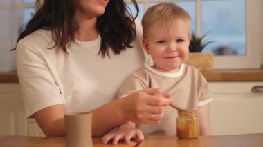 Evde mutlu bir aile. Anne, bebeğini mutfakta kaşıkla besliyor. Dağınık suratlı küçük çocuk evde sağlıklı yiyecekler yiyor. Genç kadın anne oğluna yemek veriyor.
