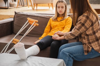 Kırık bacağı alçıda olan bir kız annesiyle evde kanepede oturuyor. Kırık bacağı iyileşen üzgün küçük çocuk oyuncak ayıya sarılıyor. Hızlı rehabilitasyon için koltuk değneklerini kullanan bir kız. Anne destek kızı