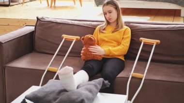 Kanepede oturan kırık bacaklı genç kız. Üzgün küçük çocuk, kırık bacağını iyileştirmek için oyuncak ayısına sarılıyor. Alçı ve koltuk değneği kullanan bir kız. Kaza sonucu oluşan kırık, ayak bandajı.