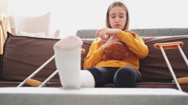 Kanepede oturan kırık bacaklı genç kız. Üzgün küçük çocuk, kırık bacağını iyileştirmek için oyuncak ayısına sarılıyor. Alçı ve koltuk değneği kullanan bir kız. Kaza sonucu oluşan kırık, ayak bandajı.