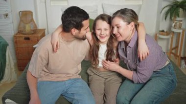 Evde mutlu bir aile. Anne, baba, kız, kanepede uzanıyor. Anne baba ve küçük kız sarılıp eğlenerek rahatlıyor. Gülümseyen aile, şefkatli anların tadını çıkarıyor